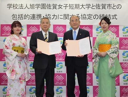 背景には両組織のロゴが並ぶパネルが設置された、学校法人旭学園佐賀女子短期大学と佐賀市の包括的連携・協力に関する協定の締結式にて、協定書を手に持つスーツ姿の男性二名と振袖姿の女性二名が並んでいる記念写真
