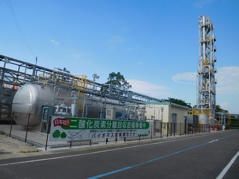 青空の下、銀色の巨大な円筒形タンクや高くそびえる蒸留塔のようなプラント設備が並び、「二酸化炭素分離回収設備稼働中」と書かれた横断幕がフェンスに掲げられている、バイオマス産業都市の施設の外観を捉えた写真