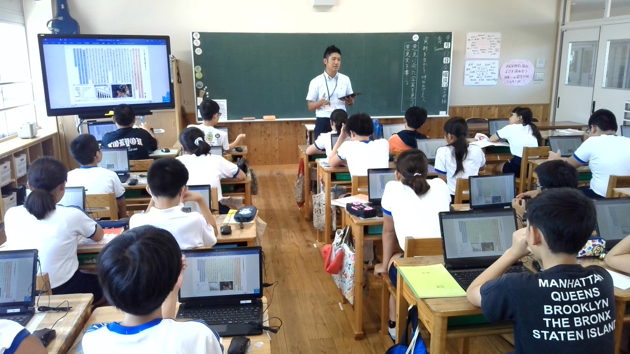 明るい教室で、黒板の前に立つ男性教師がタブレットを手に授業を行い、各自の机でノートパソコンを操作しながら大型モニターや教師に注目して学習に励んでいる児童たちの様子を背後から捉えた写真