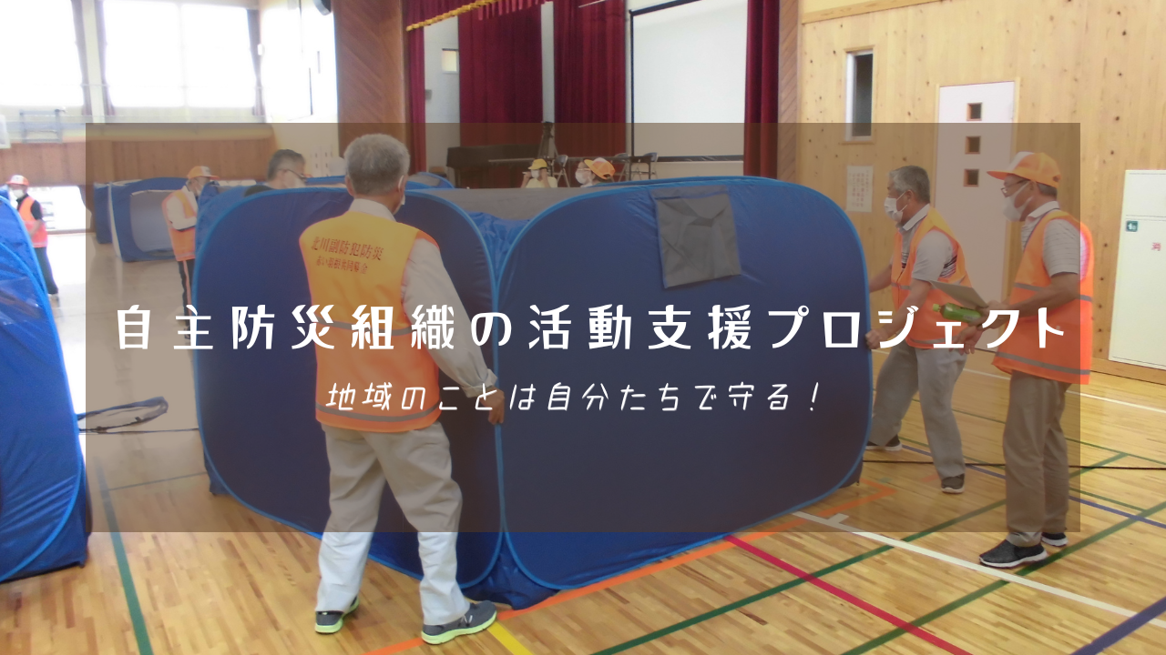 体育館でオレンジ色のベストを着用した人々が青い避難所用パーテーションを設置している様子を背景に、「自主防災組織の活動支援プロジェクト 地域のことは自分たちで守る!」という白い文字が中央に配置された写真