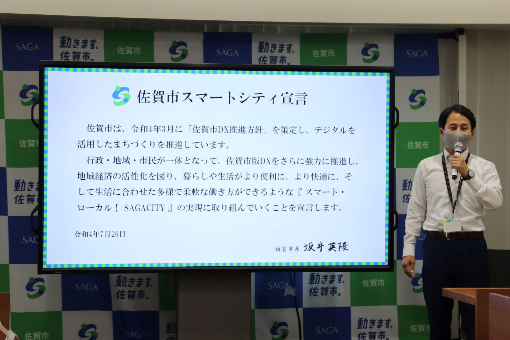 佐賀市スマートシティ宣言のテキストが映された大型モニターの横で、マイクを手にした市長がプレゼンテーションを行っており、背景には佐賀市のロゴが並ぶバックパネルが設置されている、行政の取り組みを紹介する写真