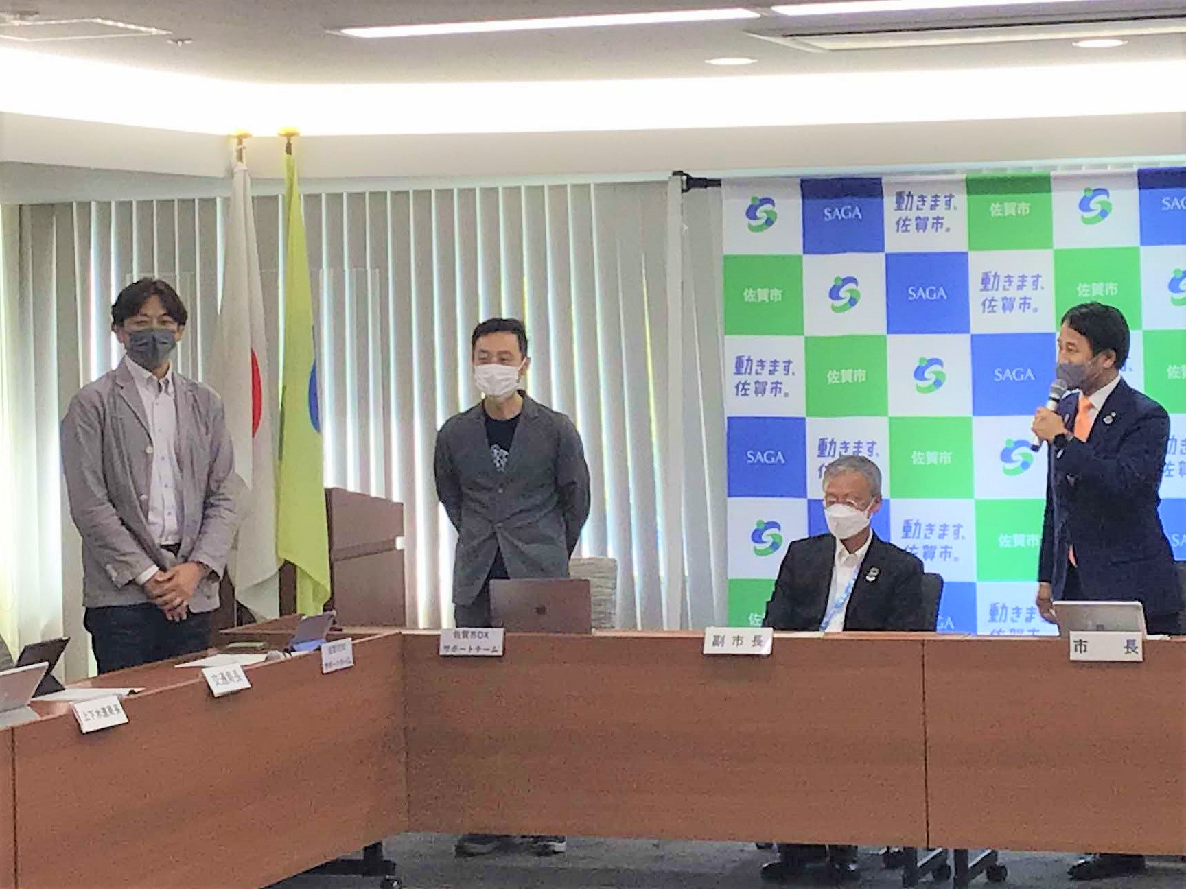 「動きます、佐賀市。」などの文字やロゴが並ぶ市松模様のバックパネルを背に、マイクを持つ市長や着席する副市長の横で、マスク姿の男性二人が立ち並び、発表を行っている様子を捉えた写真