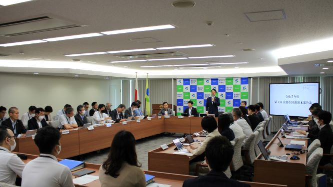 多くの参加者がコの字型の机を囲んで会議を行っており、正面のモニターには会議のタイトルが映し出され、奥では一人の男性が立って発言しており、全体的に真剣な議論が交わされている様子を捉えた室内の写真