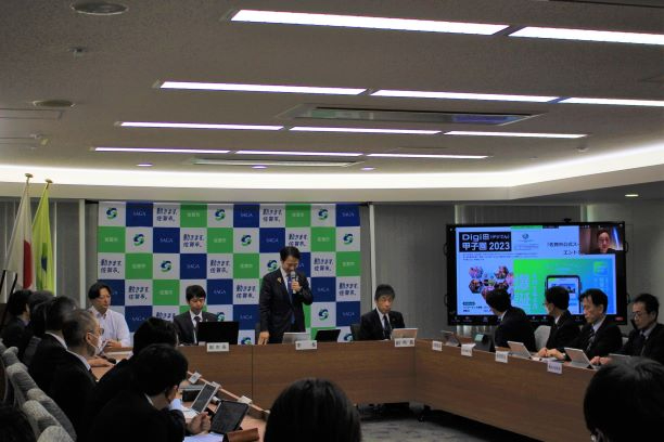 会議室で多数の参加者が着席し大型画面の発表資料を確認しながら長机を囲んで議論する様子を写した写真