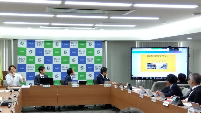 会議室で参加者たちが長机を囲み大型画面の発表資料に注目しながら議論を進める様子を写した写真