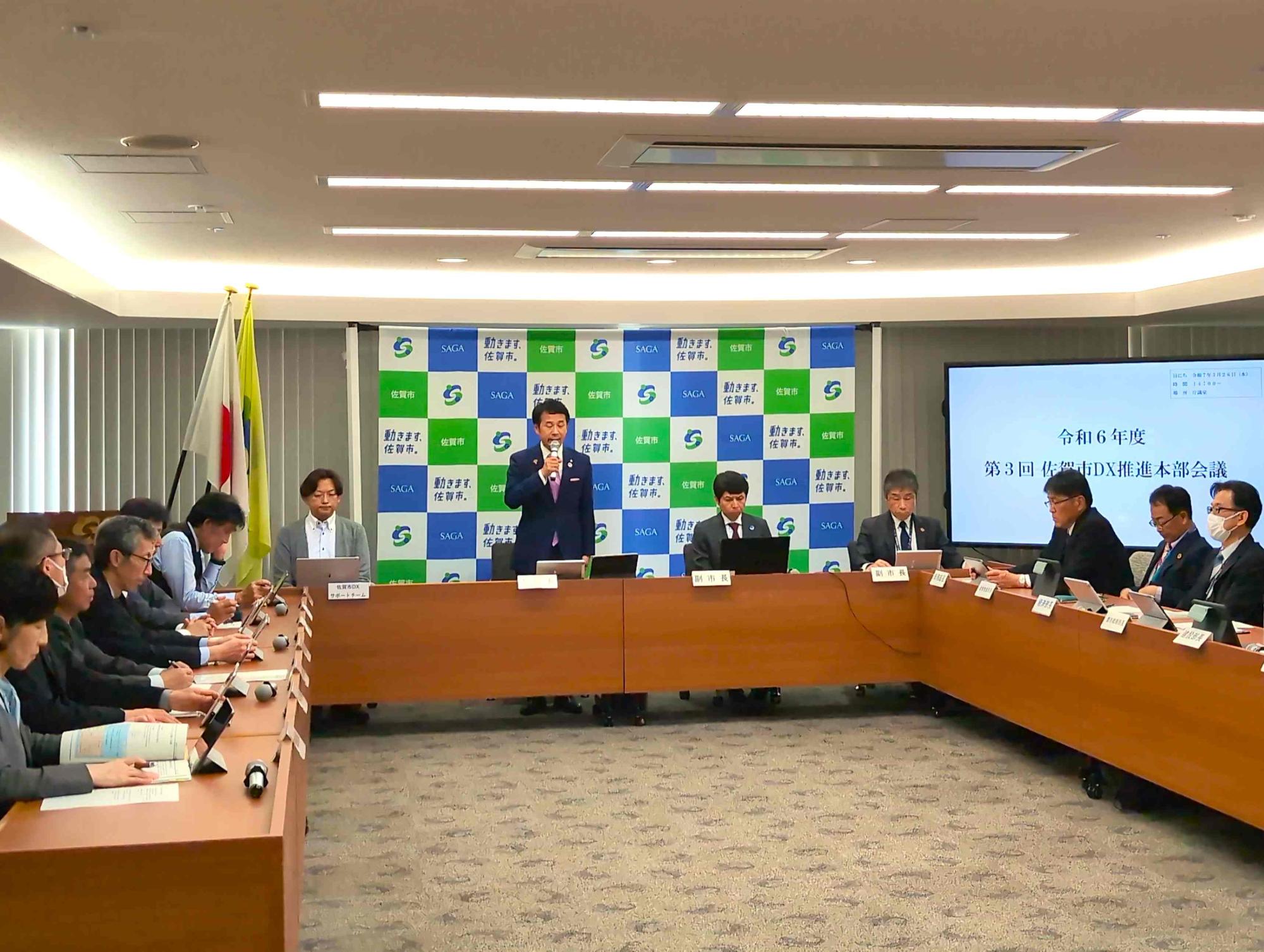 会議室で発表者が説明し参加者が長机を囲んで資料や画面を確認しながら進行する会議風景の写真