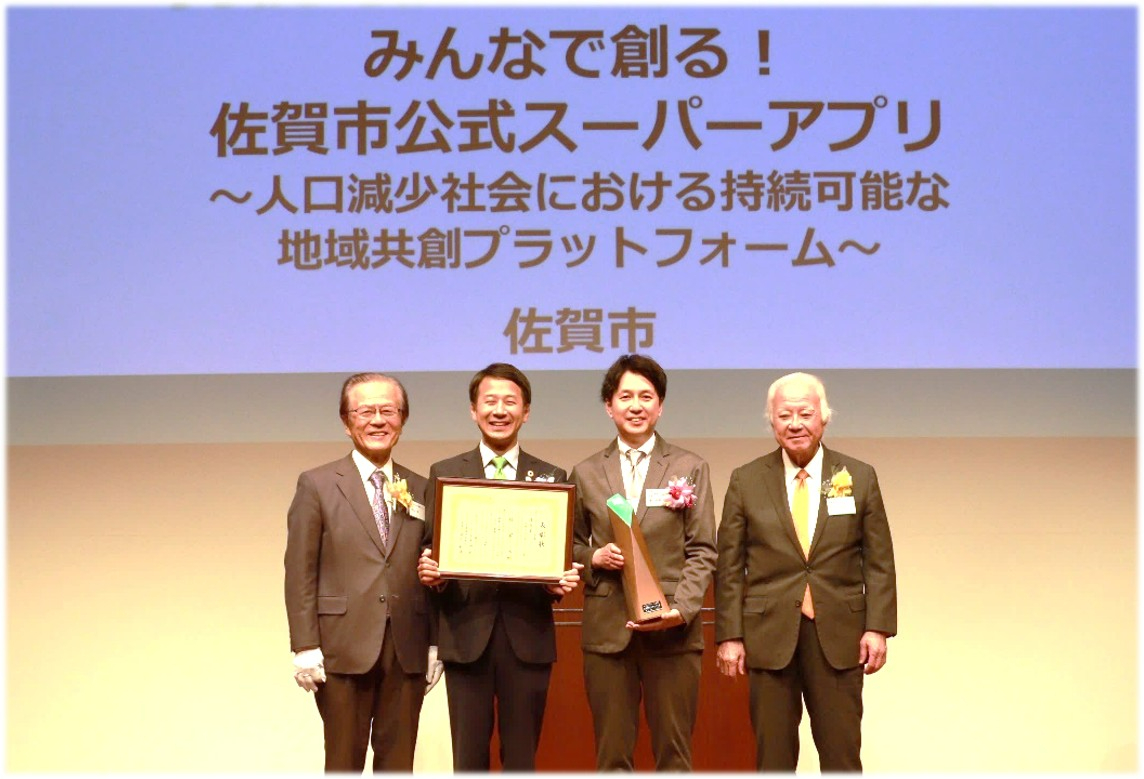 「みんなで創る!佐賀市公式スーパーアプリ〜人口減少社会における持続可能な地域共創プラットフォーム〜」と書かれたスクリーンの前で、賞状と盾を持つ男性2人を中心にスーツ姿の男性4人が笑顔で並ぶ様子の写真
