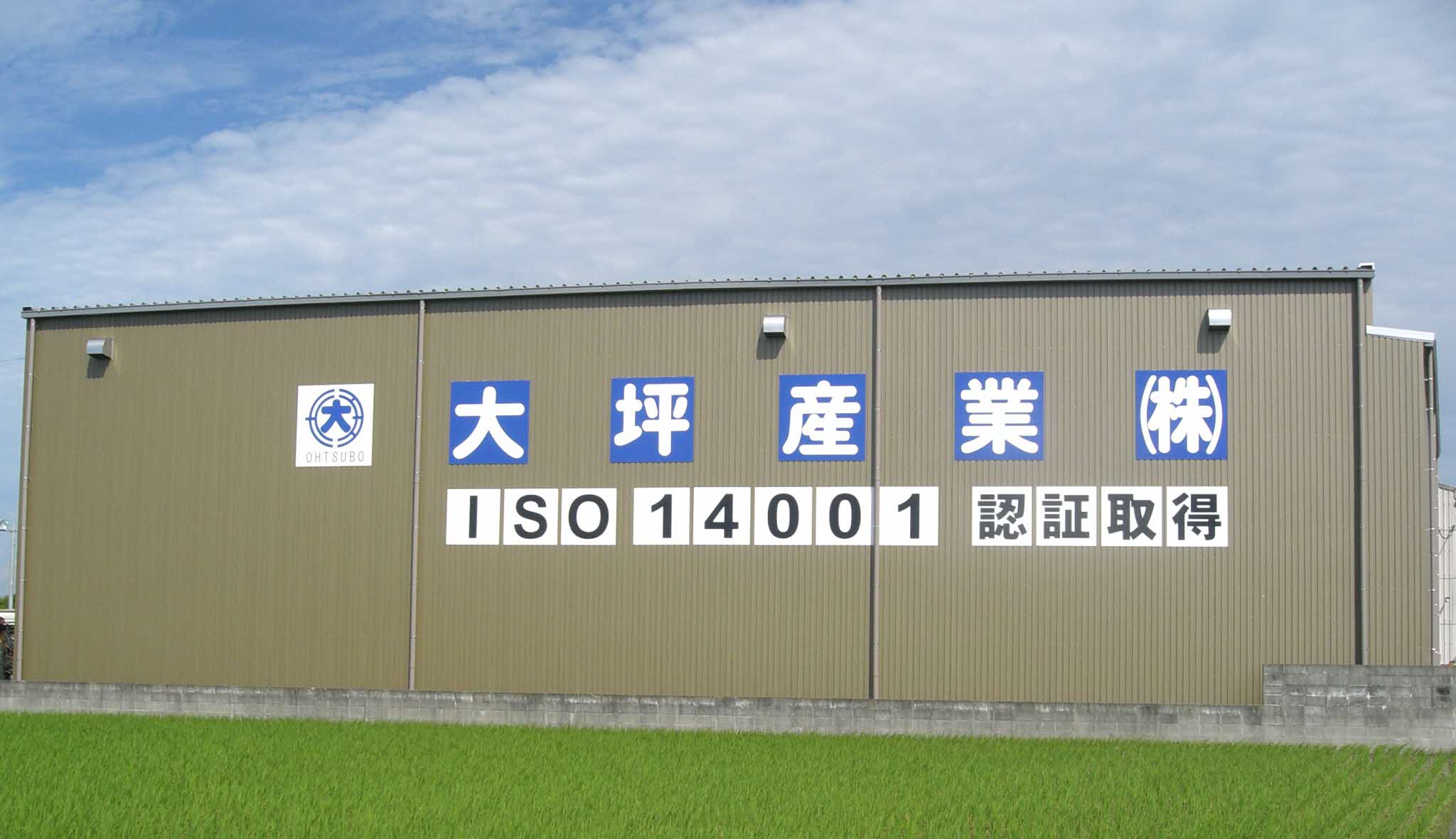 青々とした水田の奥に立つベージュ色の大きな倉庫のような建物の外壁に、青い四角枠に白抜きで「大坪産業株式会社」の社名とロゴ、その下に白地に黒文字で「ISO 14001 認証取得」と大きく掲示された、大坪産業の外観写真