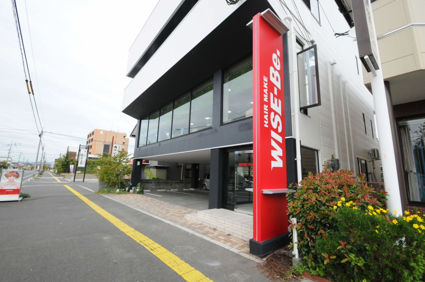 モノトーンを基調としたモダンな3階建てビルの1階に、鮮やかな赤地に白抜き文字で「HAIR MAKE WISE-Be.」と大きく書かれた縦長の自立看板が設置されているOFFICE-WISEの外観写真