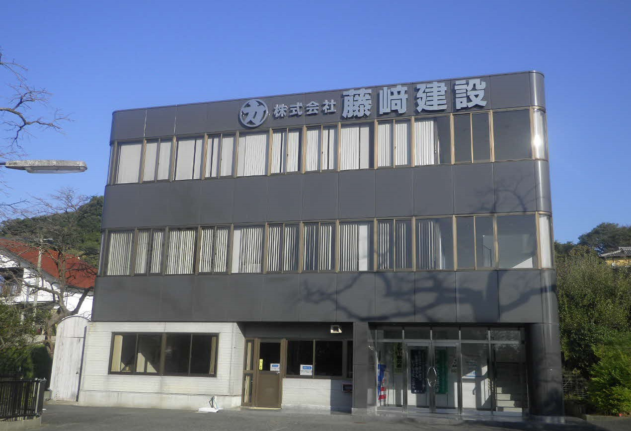 グレーのパネル貼りの外壁と大きな窓が整然と並ぶ3階建ての自社ビルで、屋上付近の壁面には丸の中に「力」の文字をあしらったロゴマークと「株式会社 藤崎建設」という社名が白く立体的に掲げられている外観写真