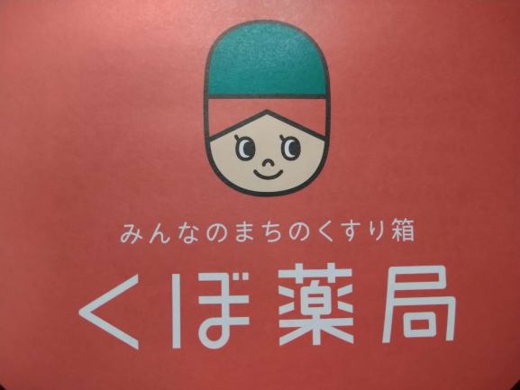 明るい赤色を背景に、緑色の帽子を被った笑顔のキャラクターの顔が中央に描かれ、その下には「みんなのまちのくすり箱」という小さなキャッチコピーと「くぼ薬局」という大きな白い文字が配置されているロゴイラスト