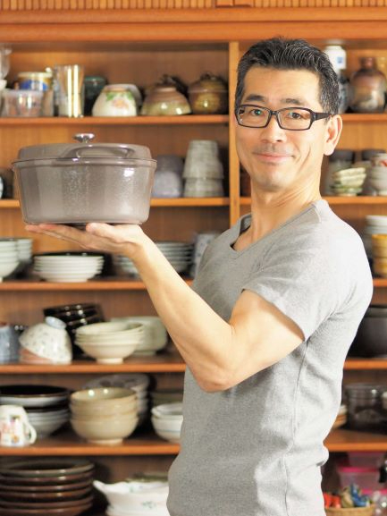 木製の食器棚に並ぶ多くの茶碗や皿を背景に、黒縁の眼鏡をかけグレーのシャツを着た男性が、左手の掌の上に蓋の付いたグレーの鋳物ホーロー鍋を載せて穏やかに微笑みながら立っている藤吉和男さんの写真。