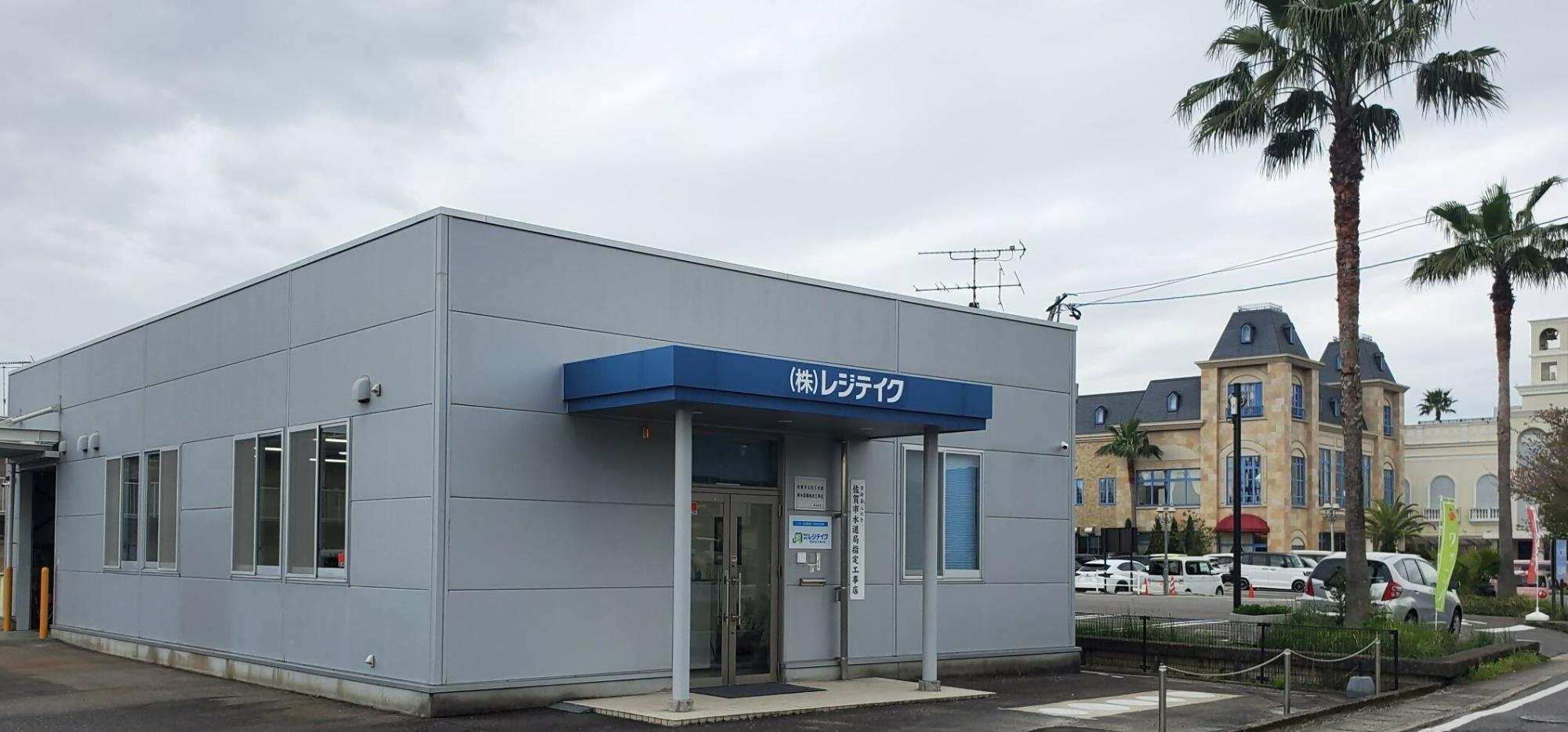 「株式会社レジテイク」という社名入りの青い庇がある平屋建てのグレーの外壁の事務所の外観写真
