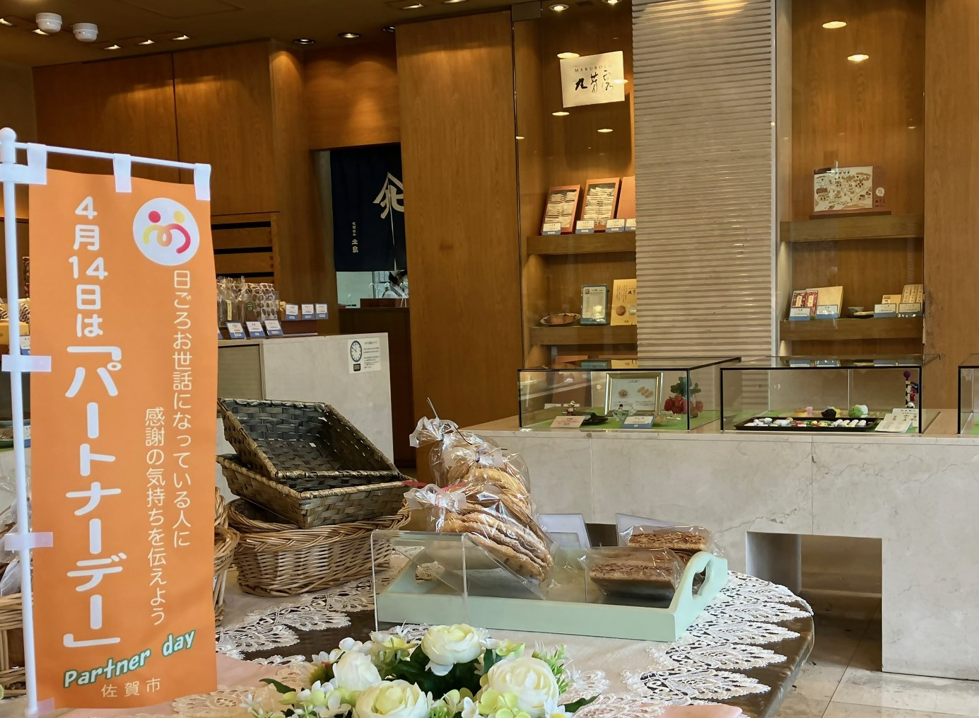 手前のテーブルに籠や個包装の菓子、奥の棚には贈答用の箱菓子が並ぶ佐賀市の和菓子店の店内に、「4月14日はパートナーデー」と書かれたオレンジ色の幟が立っている写真