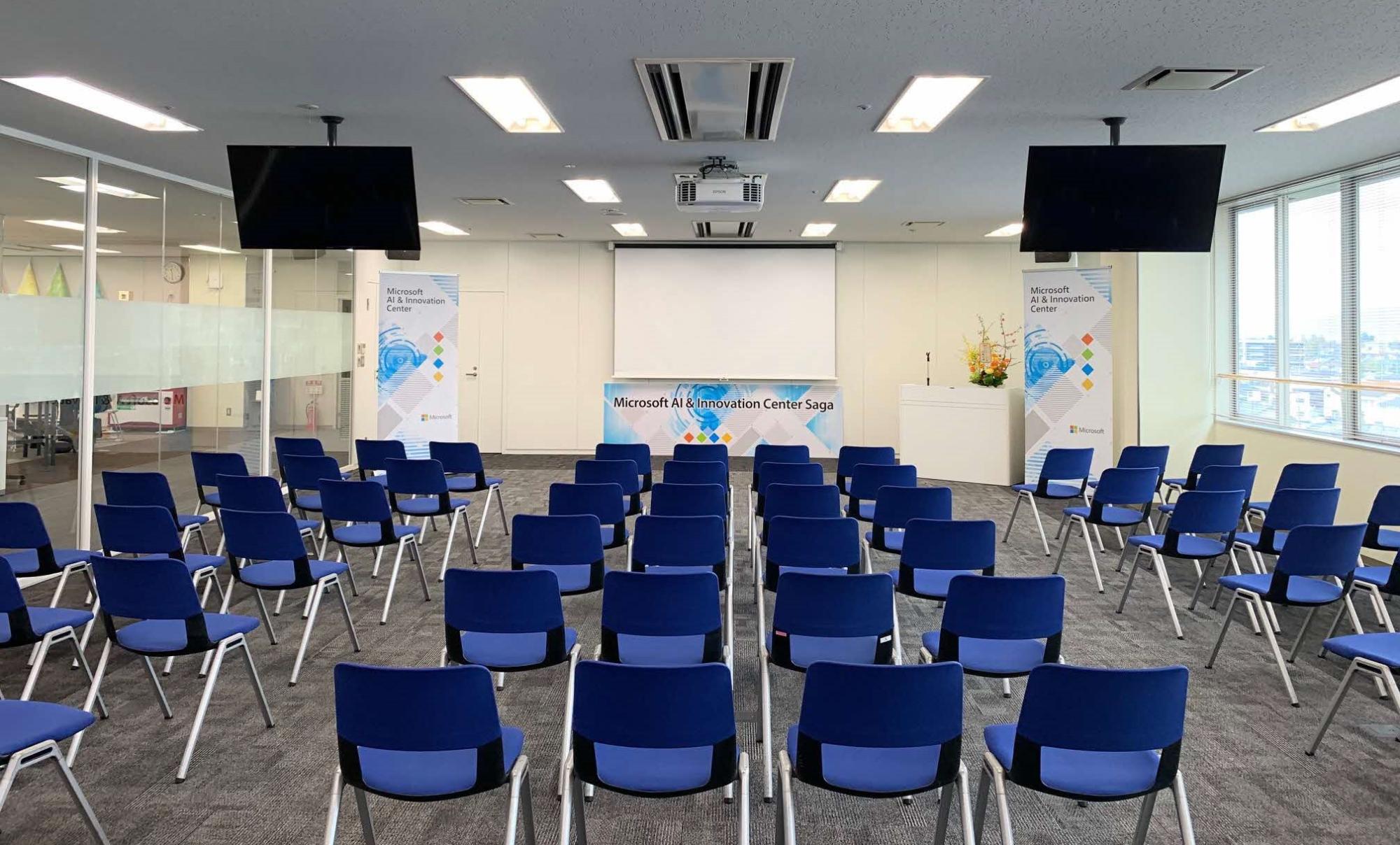 「Microsoft AI & Innovation Center Saga」のロゴが掲げられた演台とスクリーンを中心に、青い椅子が整然と並べられた、清潔感のある明るいセミナールームの全景を捉えた写真