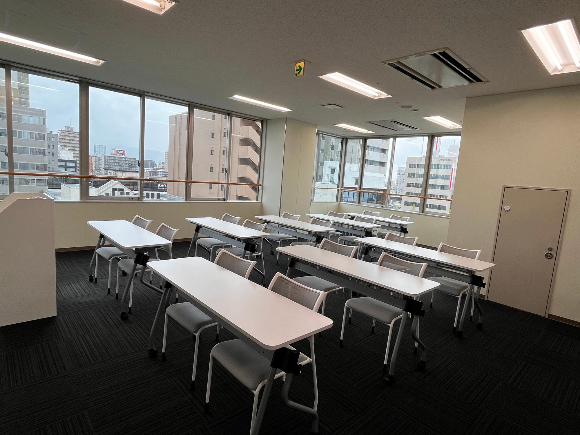 窓の外に都会のビル群が見える明るい室内で、黒いカーペットの床に白い天板のデスクとグレーの椅子が整然と並べられており、講義室や会議室として利用されるような清潔感のある広々とした空間を捉えたセミナールーム(小)の写真