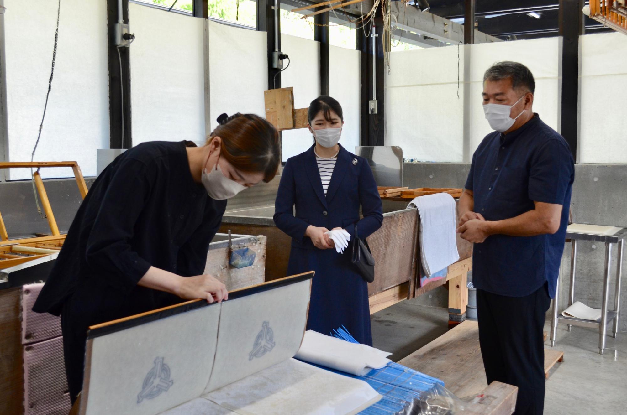 工房内で女性が和紙作りの作業に真剣な表情で取り組み、その様子を紺色のスーツ姿の愛子内親王殿下と男性が傍らで見守っている写真