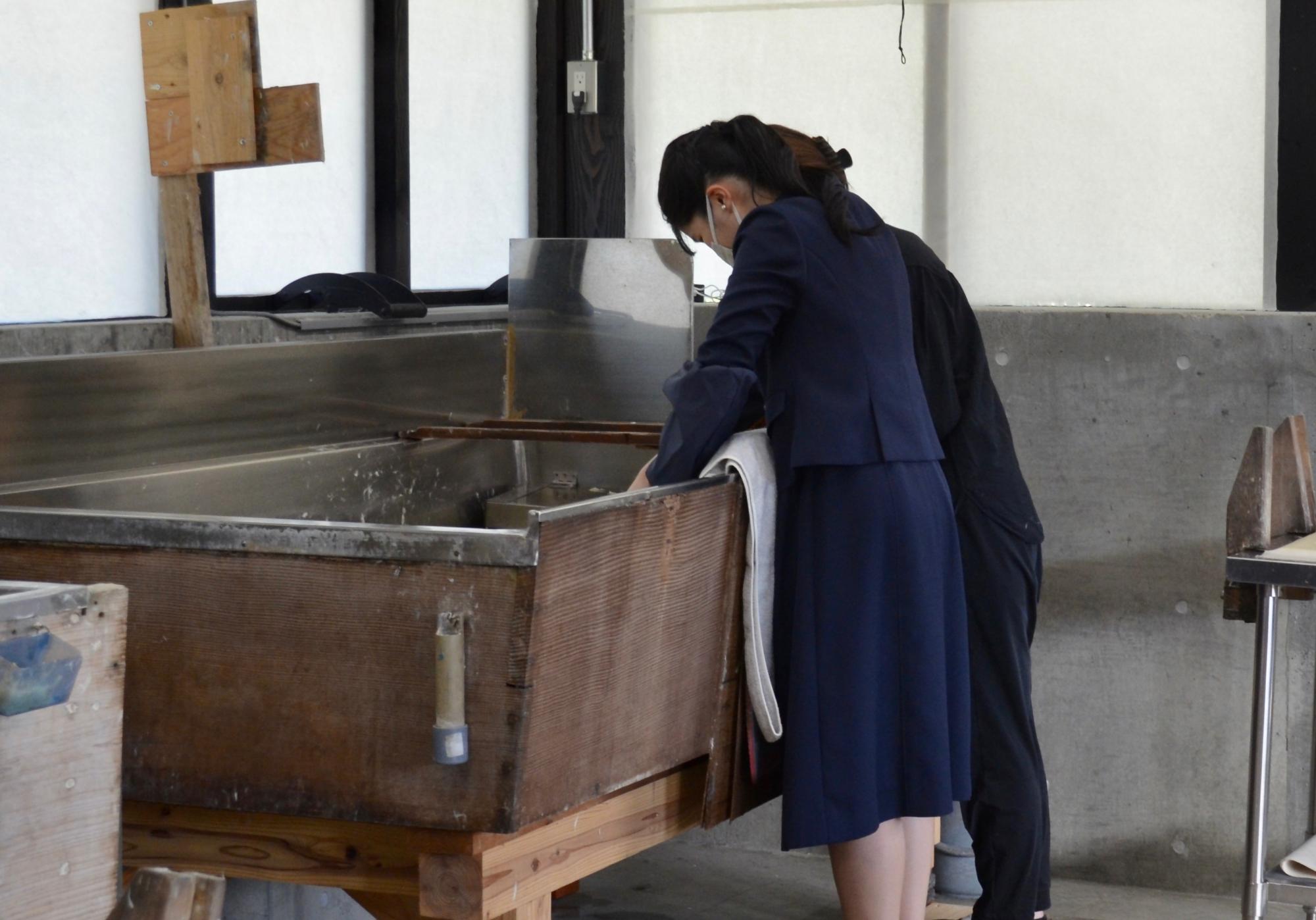 明るい工房の中で紺色のスーツを姿の愛子内親王殿下が、女性スタッフに教えてもらいながら伝統的な紙すきを体験している様子を背後から捉えた写真