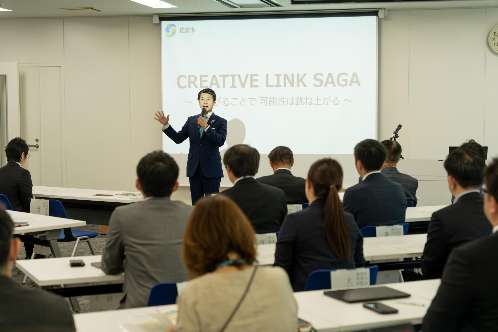 「CREATIVE LINK SAGA 〜つながることで 可能性は跳ね上がる〜」と投影されたスクリーンの前で、スーツ姿の男性がマイクを手に身振りを交えてプレゼンテーションを行い、それを見守る聴衆が背後から写されたセミナー風景の写真