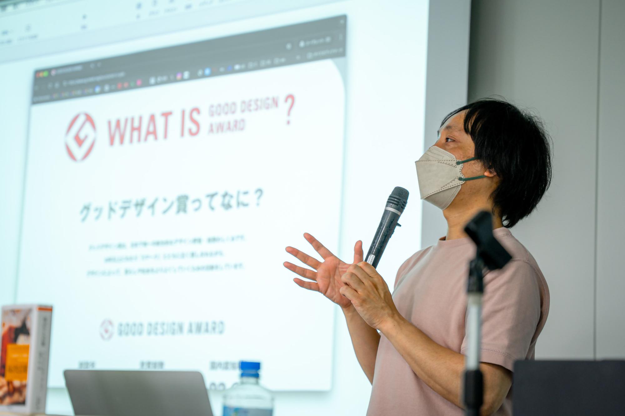 「グッドデザイン賞ってなに?」と書かれたスライドの前で、男性講師がマイクを握りながら、身振り手振りを交えて熱心にデザインの意義や賞の概要について受講生へ語りかけているセミナーの一場面を写した写真