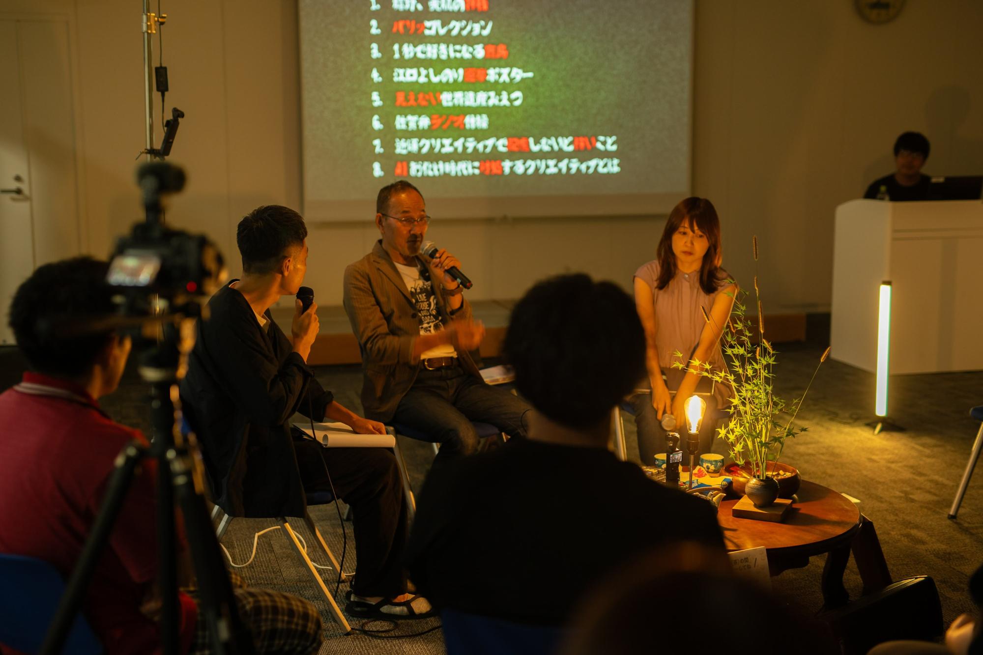 背後のスクリーンにリストが表示された薄暗い会場で、マイクを持つ男性と座っている女性のパネリストが、手前のカメラや観客に向かって対談を行っている様子を、卓上の温かい照明や植物が情緒的に演出している座談会の写真