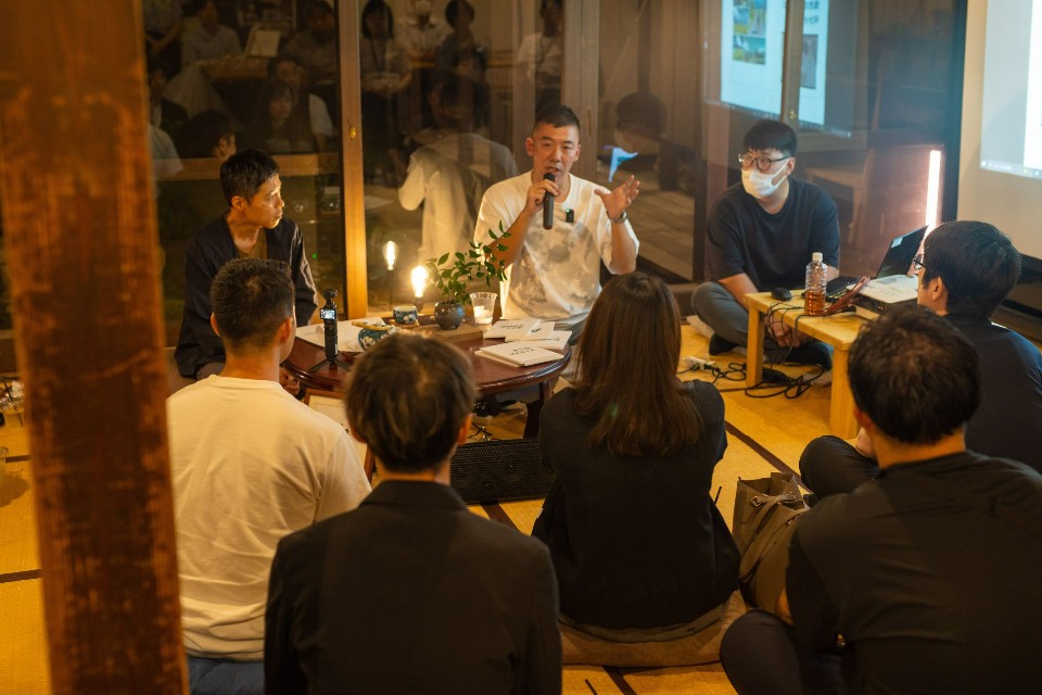 畳敷きの和室で、マイクを手に熱心に語る男性を囲むように参加者が床に座り、卓上の小さなランプや植物が置かれた円卓を囲んで親密な雰囲気の中で進められている、夜の座談会の様子を捉えた写真