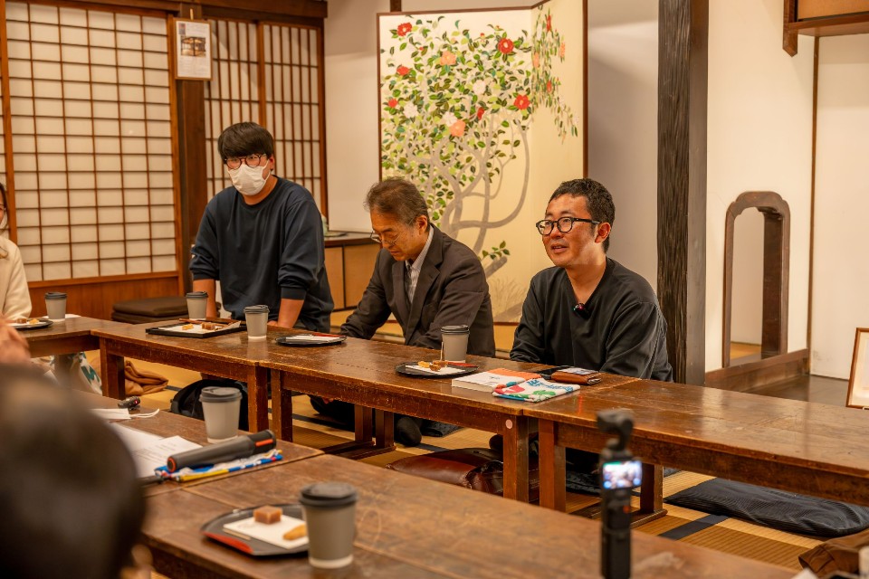 趣のある和室の長机を囲み、眼鏡をかけた男性やスーツ姿の男性たちが座ってお茶菓子を楽しみながら、落ち着いた雰囲気の中で談笑や議論を交わしている、伝統的な日本家屋で開催された座談会の様子を捉えた写真