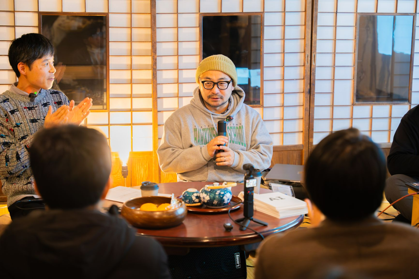 障子の前でニット帽と眼鏡を着用した男性がマイクを手に語り、隣の男性が拍手を送る様子を、茶器やみかんが置かれた円卓を囲む参加者の視点から捉えた、和気あいあいとした雰囲気の座談会の写真
