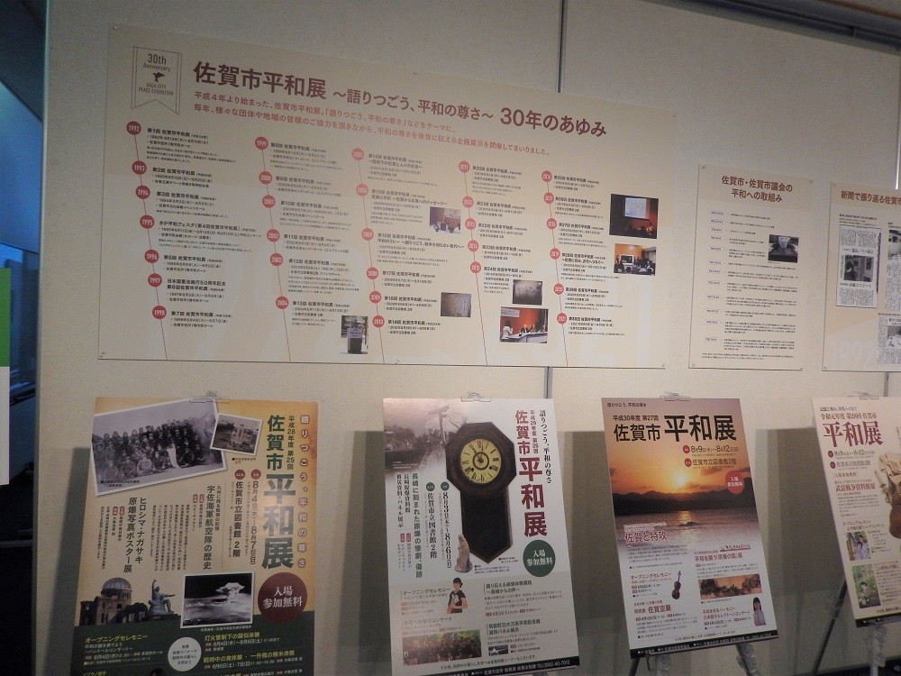 佐賀市平和展30年のあゆみと題された壁面展示パネルに歴代の平和展ポスターやチラシが年代順に配置され、下部のケースに複数のポスターやチラシの実物が陳列された記念展示の写真