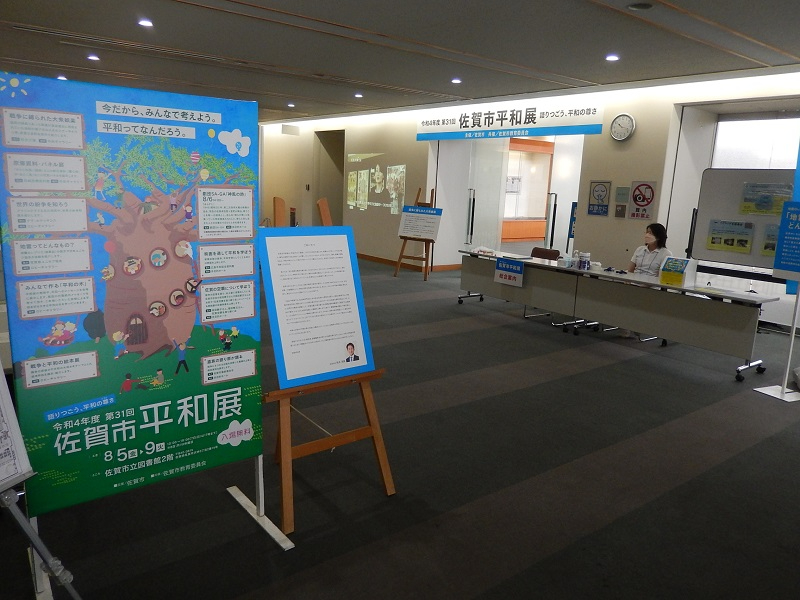 佐賀市立図書館で開催された「第31回佐賀市平和展」の会場入り口付近の様子で、手前に大きな案内ポスターやイーゼルに載った説明パネルが置かれ、奥には受付デスクと座っているスタッフ、展示室への通路が見える写真