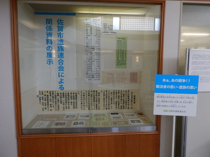 ガラス張りの展示ケース内に「佐賀市遺族連合会による関係資料の展示」という看板が掲出され、戦地別の戦没者数一覧や軍隊手帳の説明パネル、底部には当時の写真や手紙などの貴重な遺品が整然と並べられた展示風景の写真