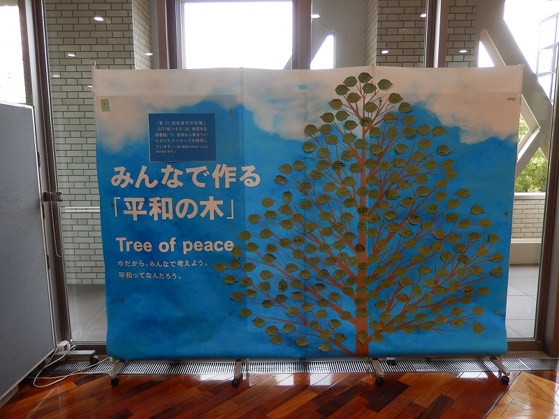 青空を背景に大きな木のイラストが描かれた「みんなで作る平和の木」という掲示パネルがあり、木の葉に見立てた多数のメッセージカードが貼られ、平和への願いや考えを共有する参加型展示の様子を正面から捉えた写真