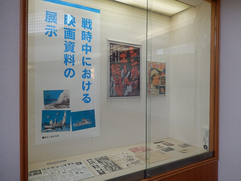 ガラス張りの展示ケース内に「戦時中における映画資料の展示」と書かれた青い縦長のポスターが貼られ、その横に当時の映画のチラシや、ケース底面には新聞の切り抜きなどの貴重な紙資料が丁寧に並べられた展示風景の写真