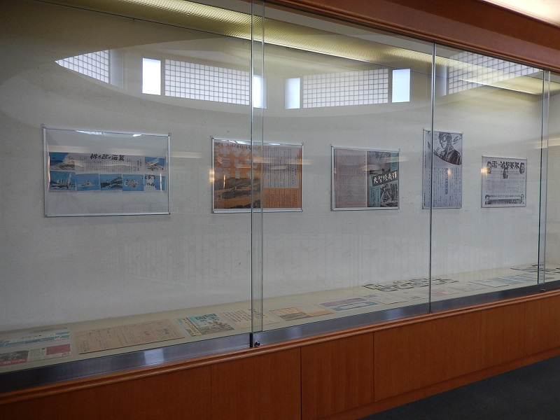 ガラス張りの大きな展示ケース内に、当時の新聞記事や写真パネルが等間隔で掲示され、手前の平置きスペースには古い冊子や切り抜き資料が並べられており、背後のガラスには吹き抜け天井の採光窓が映り込んでいる写真