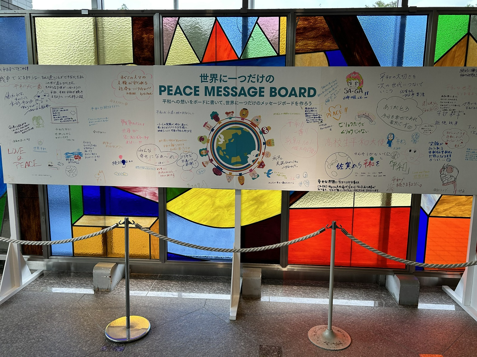 カラフルなステンドグラスを背景に、「世界に一つだけの PEACE MESSAGE BOARD」と記された大きな白いボードが設置され、来場者による平和への願いやイラストが自由に書き込まれている様子を正面から捉えた写真