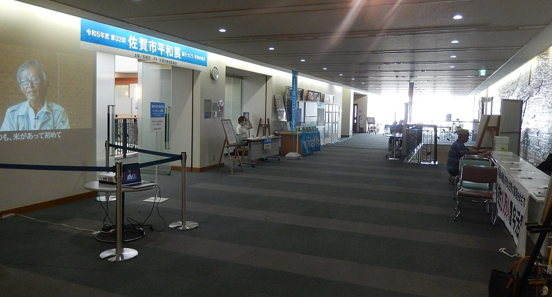 「令和5年度 第32回 佐賀市平和展」と書かれた青い横断幕が掲げられた屋内の展示会場にて、壁面にビデオ映像が投影され、奥にはユニセフの旗や受付デスク並べられた写真