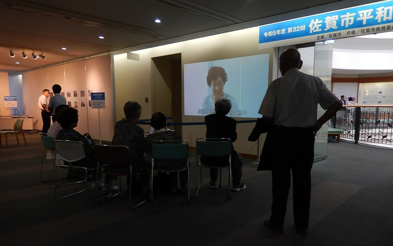 「令和5年度 第32回 佐賀市平和展」と書かれた青い看板の下で、大型スクリーンに映し出された戦争体験者の証言映像を、パイプ椅子に座った数名の来場者が視聴している写真