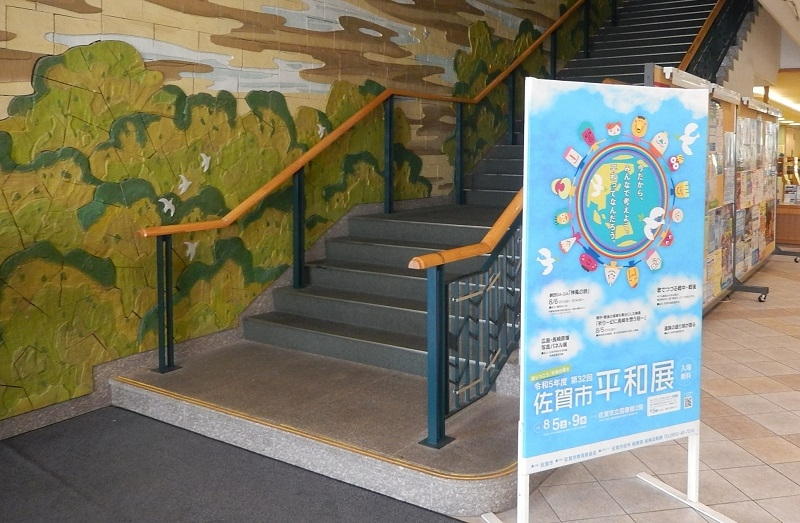 「令和5年度 第32回 佐賀市平和展」の開催を告げる水色のポスターが手前に配置され、背景には緑豊かな風景が描かれたレリーフ状の壁面と、上階へと続く階段がある屋内ロビーの写真