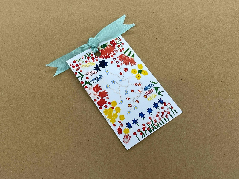 長方形の白いカードに赤や黄色、青といった色鮮やかな花々が手書き風のタッチで描かれ、上部には淡いミントグリーンのリボンが結ばれているしおりの写真
