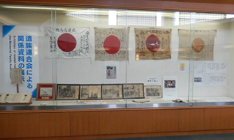 ガラス張りの展示ケース内に、「遺族連合会による関係資料の展示」と書かれ、寄せ書きが記された日章旗や当時の様子を伝えるモノクロ写真、資料などが整然と並べられている写真