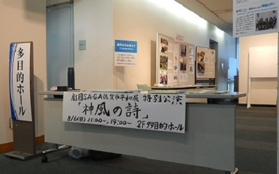 「劇団SA-GA佐賀市平和展 特別公演『神風の詩』」と手書きされた案内掲示がデスクの前面に貼られた、公演会場の受付付近を写した写真