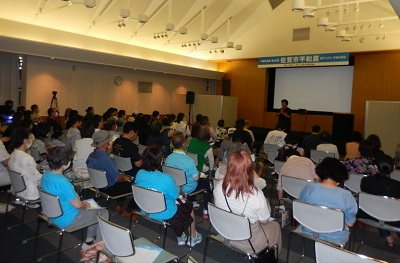 「令和5年度 第32回 佐賀市平和展」と書かれた青い横断幕が掲げられた多目的ホールのステージ前で、多数の来場者がパイプ椅子に座り、前方のスクリーンや登壇者の方を向きながら静かに耳を傾けている様子を捉えた写真