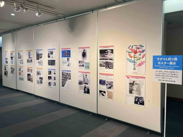 「サダコと折り鶴ポスター展示」という案内板の横で、白い展示壁に沿ってサダコさんの生涯や原爆の被害を伝える多数のパネルが並んでいる写真