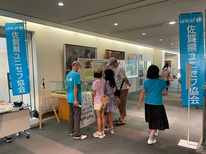 「佐賀県ユニセフ協会」と書かれた青い幟が立つ会場にて、ユニセフのTシャツを着たスタッフが、熱心に展示品を見つめる親子連れに説明を行っている写真