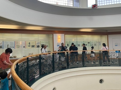 円形の吹き抜けがある明るい展示室にて、ガラスケース内に並んだパネル資料を熱心に鑑賞する複数の来場者の様子を捉えた写真