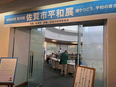 「令和6年度 第33回 佐賀市平和展」と書かれた青い横断幕が掲げられた会場入口で、数名の来場者が展示資料を熱心に見ている写真