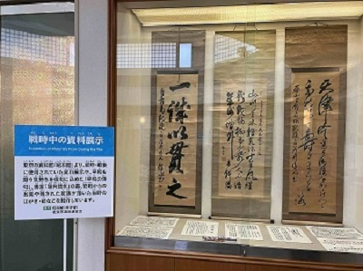 「戦時中の資料展示」という案内板の横で、ガラスケース内に飾られた三幅の古風な掛け軸、その下に並べられた当時の文書などを展示した写真