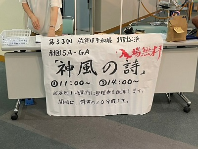 「第33回 佐賀市平和展 特別公演」として、劇団SA-GAによる「神風の詩」の公演案内が手書きで大きく記され、入場無料の赤い文字や開演時間などが書かれた模造紙が事務机の前に掲示されている会場受付の写真