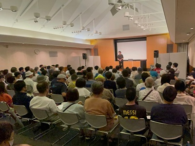 前方にスクリーンが設置されたホールにて、多くの観客が椅子に座ってステージ上の登壇者の話を熱心に聴講している会場後方から写した写真