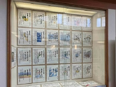 明るい照明が灯る大きな展示ケースの中に、色鮮やかな挿絵と言葉が添えられた数多くの色紙が、縦四段、横六列に整然と並べられている写真